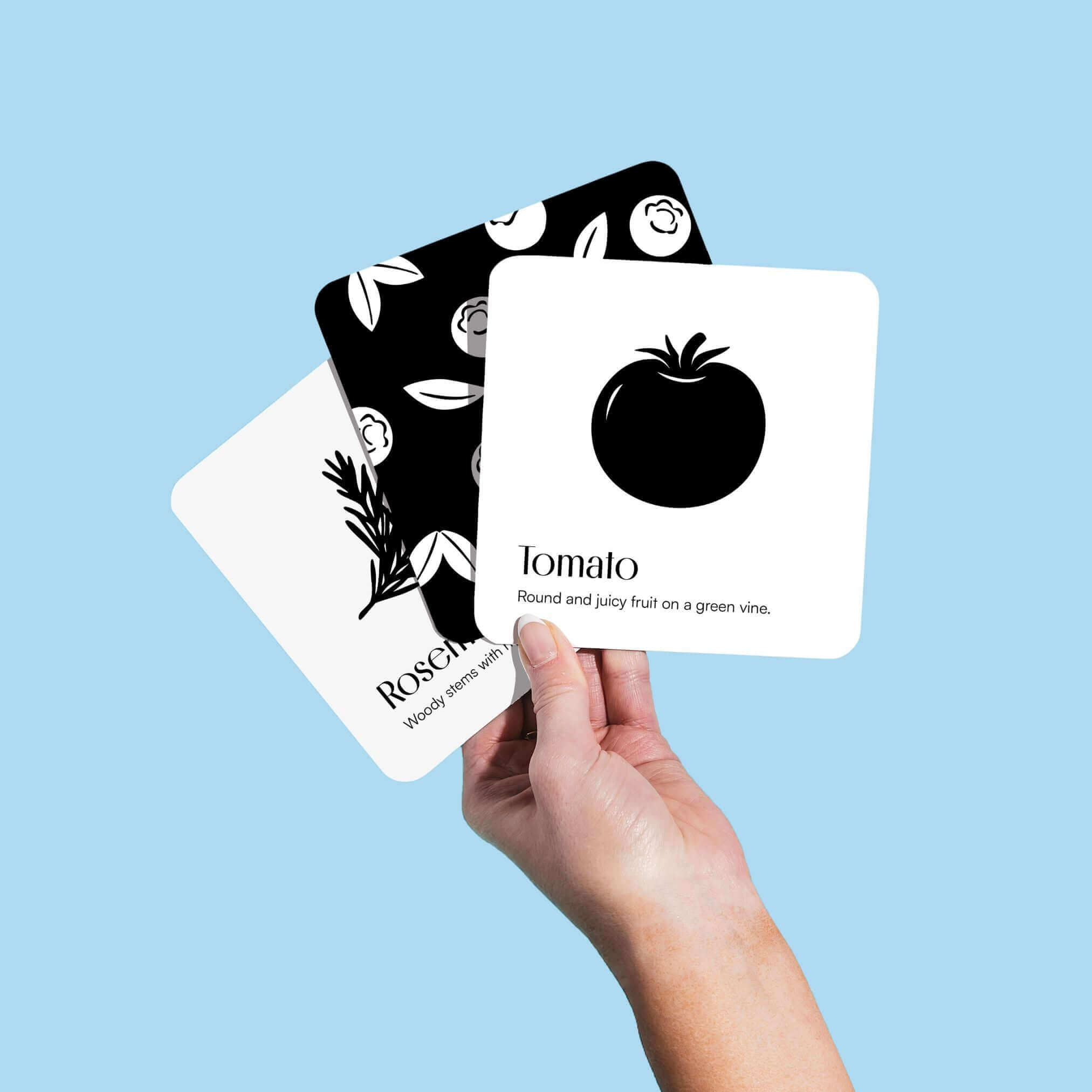 A white hand holds up three black and white "Garden for Baby" art cards against a solid light blue background. The card on top shows a tomato, the one beneath it partially shows rosemary, and the one at the back shows a pattern of flowers.