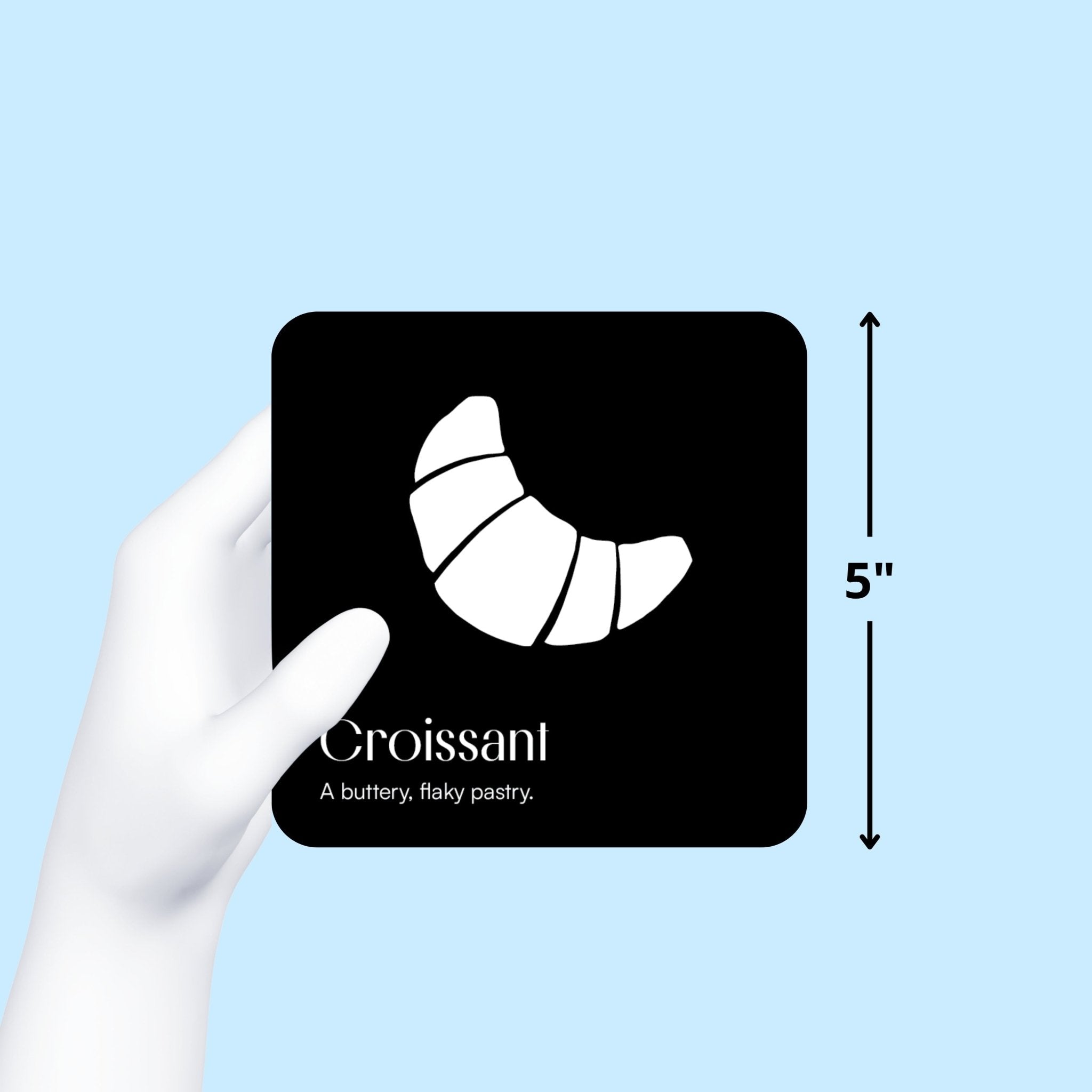 Gloved hand holds a black and white "Croissant" art card, showing its 5-inch size on a blue background from the Gourmet for Baby art cards from Chunky Deli.