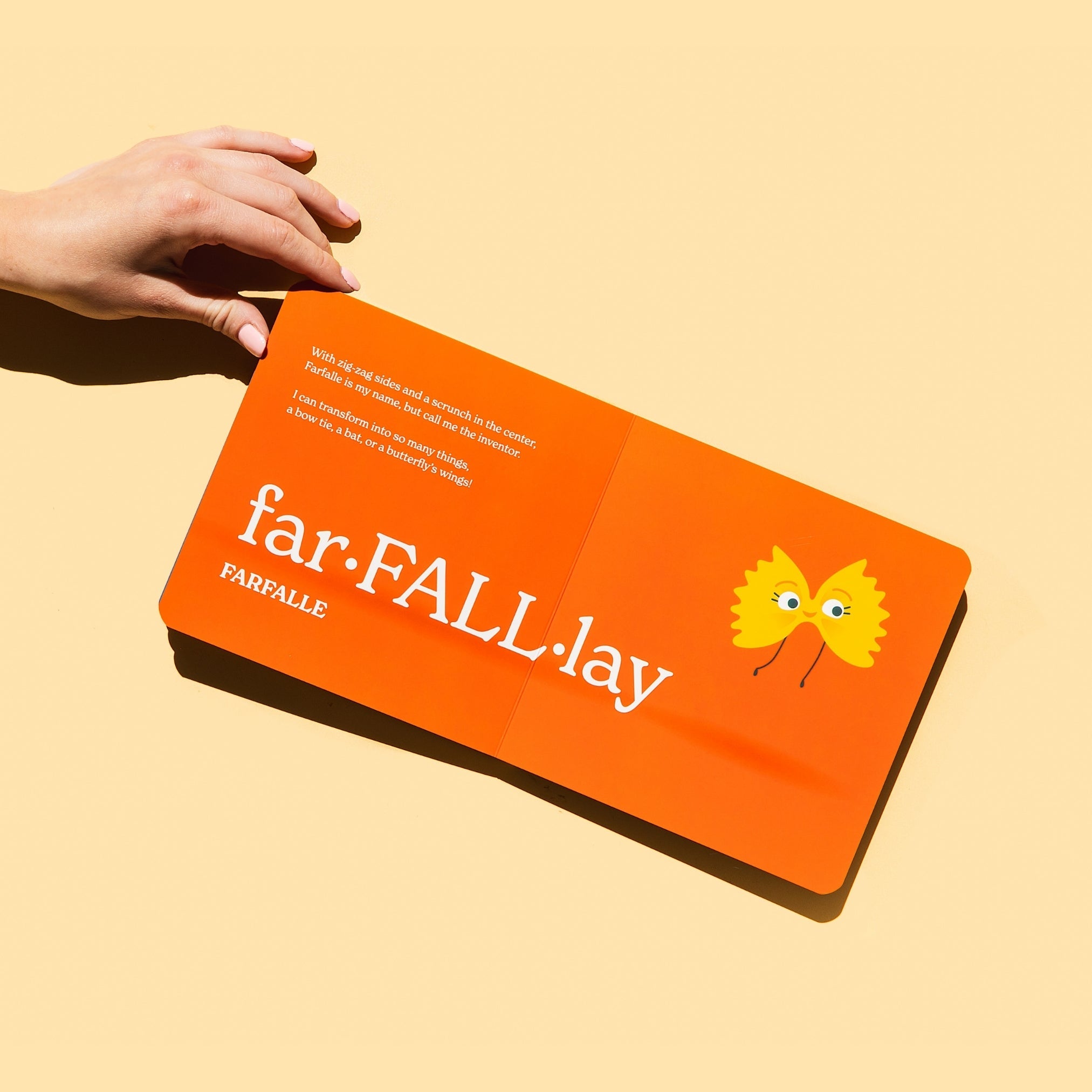 Chunky Deli product image: "Little Book of Pasta" open to the farfalle page (orange spread), with a hand holding the corner of the page, resting on a light orange background.
