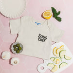 Chunky Deli product image: "Sous Chef" bodysuit folded in half and laid on a pink background, surrounded by real food items in a playful arrangement.