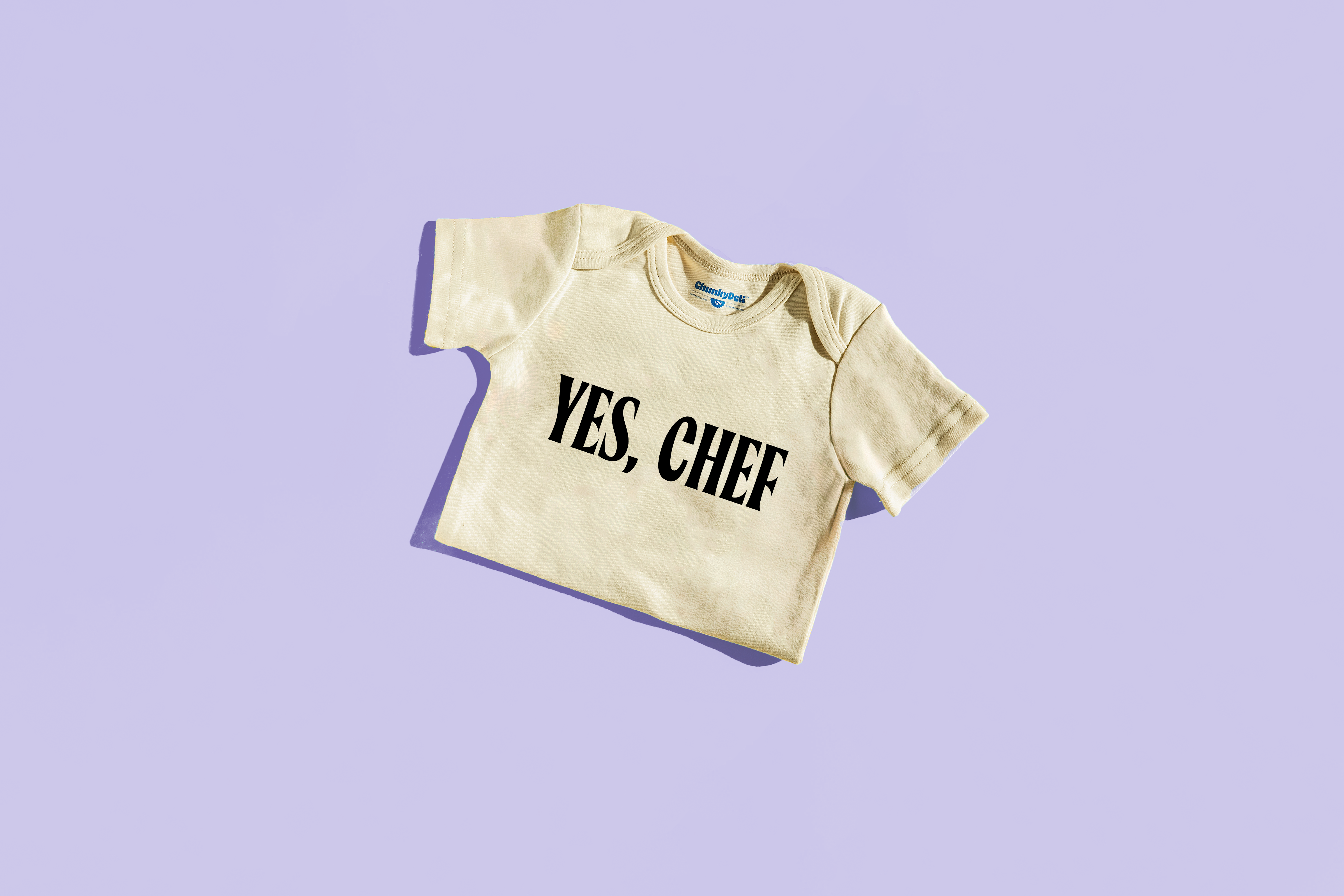 A neutral-colored Toddler Tee from Chunky Deli with the words "YES, CHEF" written in black text is shown on a light purple background.