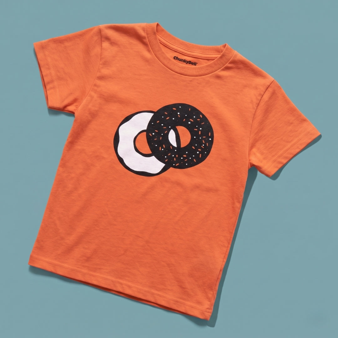 Chunky Deli product image: Toddler tee in 100% cotton, short sleeve, orange color. Features a graphic illustration of everything bagel with cream cheese in 2 colors, black and white, centered on the chest on a blue background.