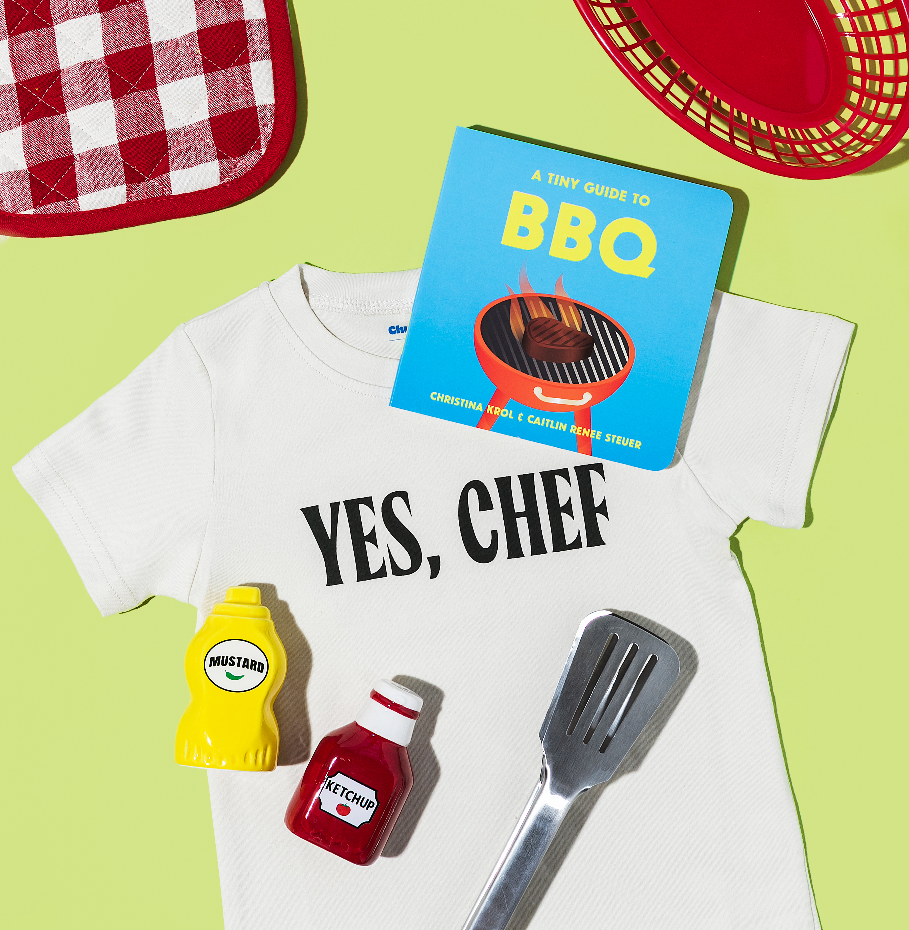 A white onesie reads "YES, CHEF" with "A tiny guide to BBQ" book, mini ketchup and mustard bottles, and a mini spatula on a green background.