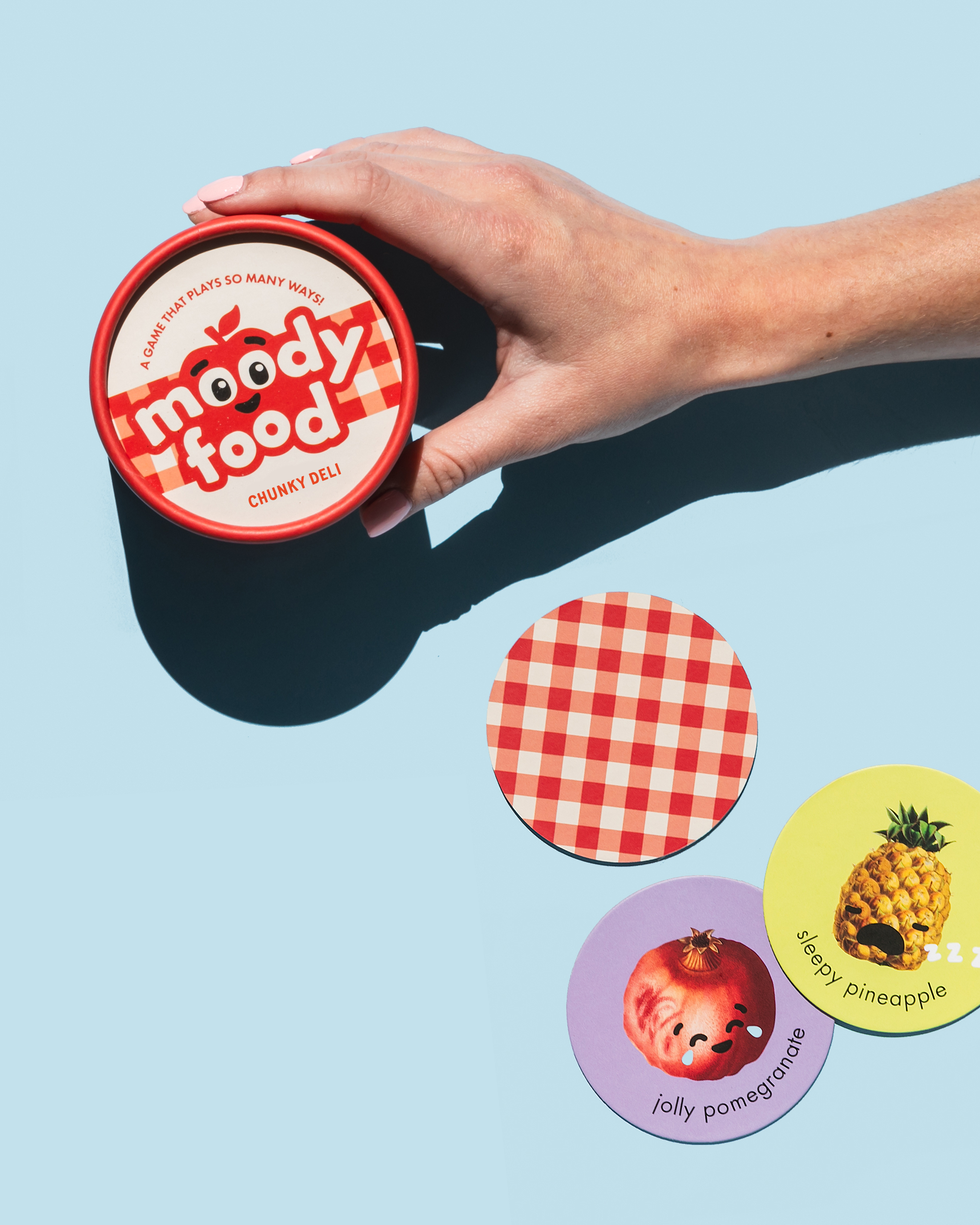 A hand holds a red and white circular box for the Chunky Deli "Moody Foody" card game. Three round cards are fanned out below it on a blue background, each with a different illustrated "moody food."
