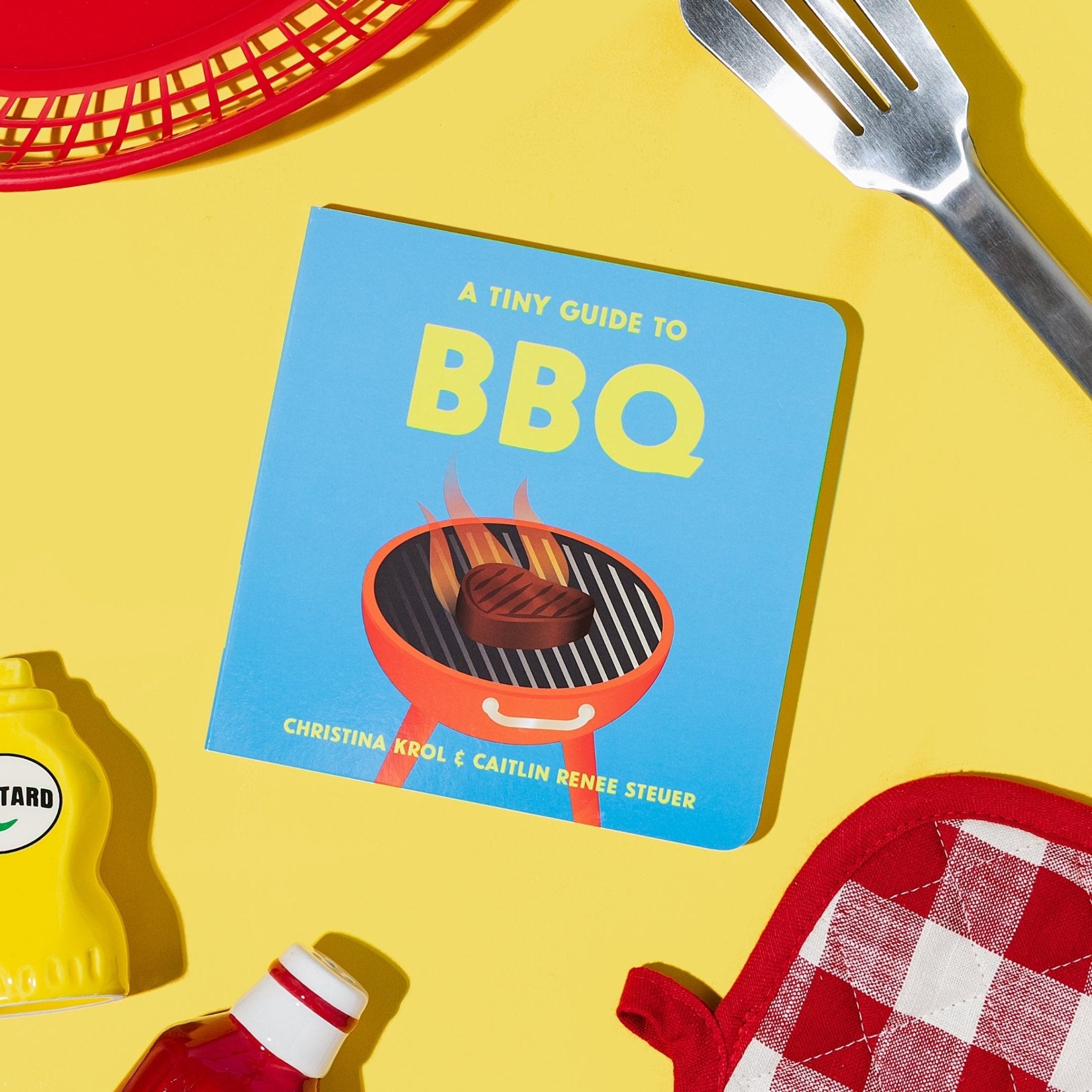 A Tiny Guide to BBQ book cover, on a yellow background with a red basket, mustard, ketchup, grill spatula, and oven mitt. From Chunky Deli Written by Christina Krol & Caitlin Renee Steuer