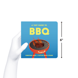 Chunky Deli product Image. The bright blue cover of the board book 'A Tiny Guide to BBQ' showcases a bold yellow title above a classic red grill with cartoon flames and a steak. Authors Caitlin Renee Steuer and Christina Krol.