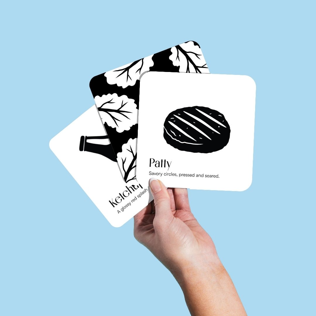 Hand holding three Chunky Deli black and white baby art cards featuring a patty and ketchup against a light blue background.