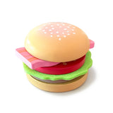 multi-layered toy hamburger from the Chunky Deli range. The stack, from top to bottom, features a light-brown wooden bun top with painted white sesame seeds, a wavy green felt lettuce leaf, a thick red wooden tomato slice, a rectangular pink wooden block representing bacon with dark stripes, a dark-brown wooden meat patty, and a light-brown wooden bun base