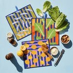 Colorful kitchen towels with fish, lemon, and leaf patterns on a blue background, surrounded with ingredients to make caesar salad (Anchovy, lemon, parmesan, croutons)