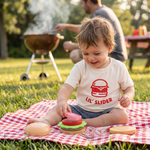 Child playing with toy burgers on a picnic blanket outdoors wearing Lil' Slider Kids' Tee from Chunky Deli.