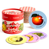 Chunky Deli product image: "Moody Food" card game, featuring a cylinder-shaped game box in the background and a selection of round cards laid in front, showcasing vintage food illustrations with emotions on a checkered background. An apple and an orange c