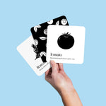  A white hand holds up three black and white "Garden for Baby" art cards against a solid light blue background. The card on top shows a tomato, the one beneath it partially shows rosemary, and the one at the back shows a pattern of flowers.