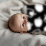 Newborn baby lies on a cream-colored blanket, with large gray eyes looking at a blurry black and white Garden for baby art card.