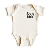 Chunky Deli product image: Natural-colored "Sous Chef" bodysuit (100% cotton) laid flat on a white background, showcasing the envelope shoulders and three-snap closure.