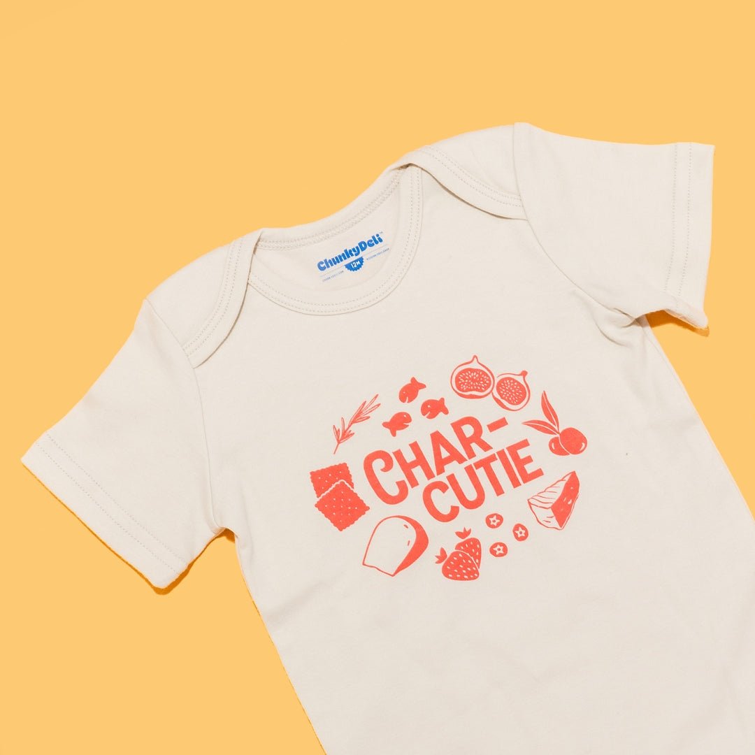  Close-up of the "Charcutie" baby bodysuit in natural cotton on an orange background. The graphic features the word "Charcutie" surrounded by food illustrations. Chunky Deli product image.