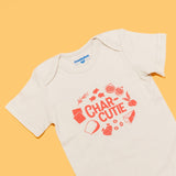  Close-up of the "Charcutie" baby bodysuit in natural cotton on an orange background. The graphic features the word "Charcutie" surrounded by food illustrations. Chunky Deli product image.