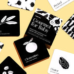 Top-down flat lay of the "Gourmet for Baby" art cards, with the box in the center, on a pastel yellow surface.
