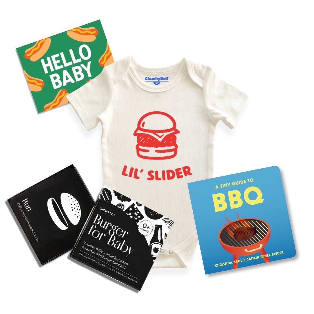 Chunky deli product image of the grillmaster baby bundle, top down view of a baby onesie with a bold graphic and "lil' Slider" Printed in orange font. There is also a tiny guide to BBQ, Burger for Baby art cards and a "Hello Baby" gift note. 