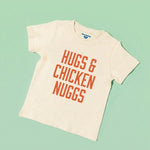 Chunky Deli toddler tee in 100% cotton, short sleeve, beige/natural color with bold orange block text reading 'Hugs & Chicken Nuggs' across the chest on a light green background. Playful foodie apparel for kids.