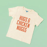 Chunky Deli toddler tee in 100% cotton, short sleeve, beige/natural color with bold orange block text reading 'Hugs & Chicken Nuggs' across the chest on a light green background. Playful foodie apparel for kids.