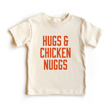Chunky Deli toddler tee in 100% cotton, short sleeve, beige/natural color with bold orange block text reading 'Hugs & Chicken Nuggs' across the chest. Playful foodie apparel for kids.