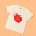 A soft white toddler t-shirt featuring a vibrant red tomato graphic and the words La Dolce Vita in elegant script on a pastel orange background. Chunky Deli Product Image