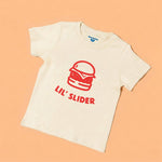 Chunky Deli product image: Toddler tee in 100% cotton, short sleeve, beige/natural color. Features a graphic illustration of a burger with the text "Lil' Slider" underneath, centered on the chest on a pastel orange background.