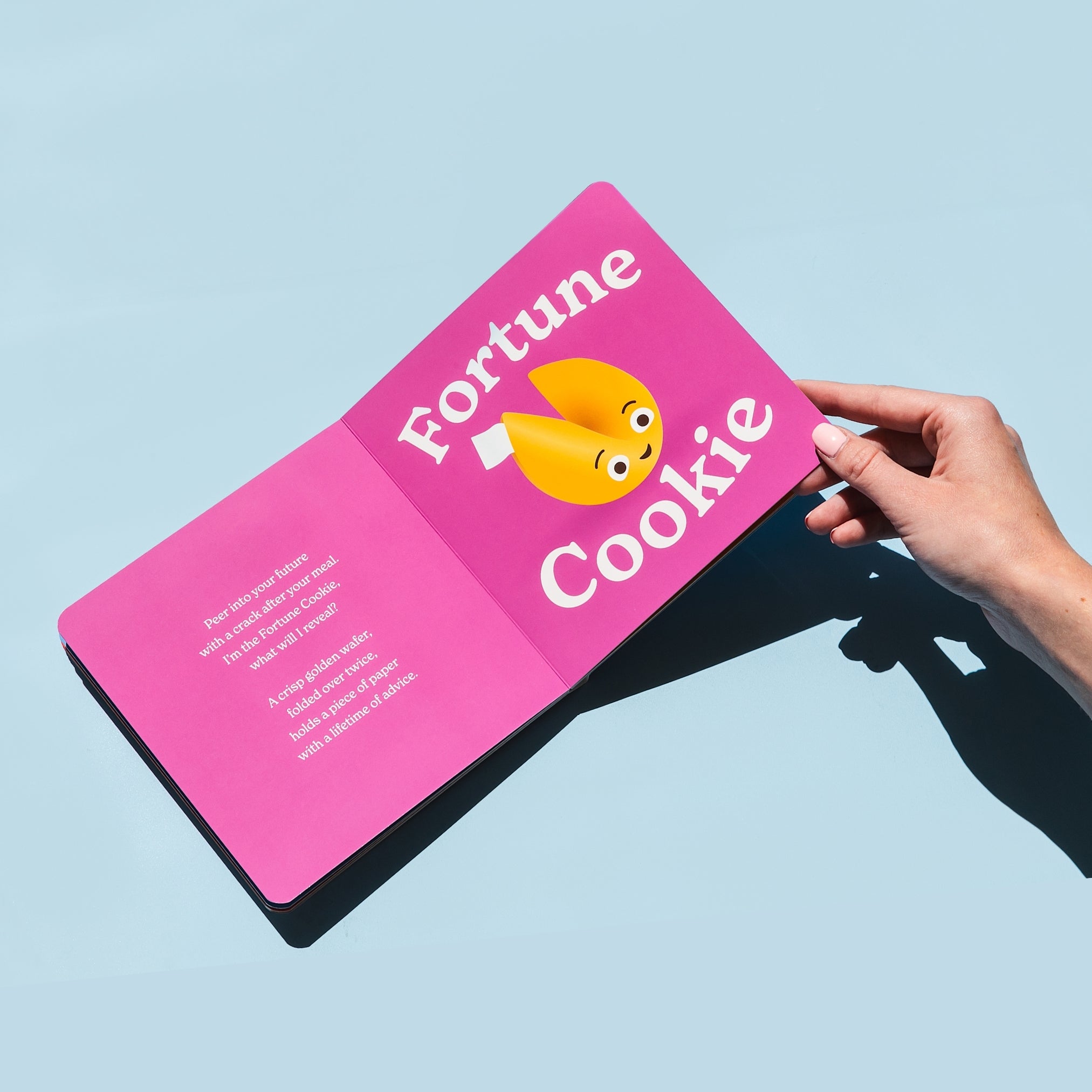 Chunky Deli product image: "Little Book of Cookies" open to the fortune cookie spread (purple page) featuring an illustration of a fortune cookie and accompanying rhyme, held open by one hand against a light blue background.