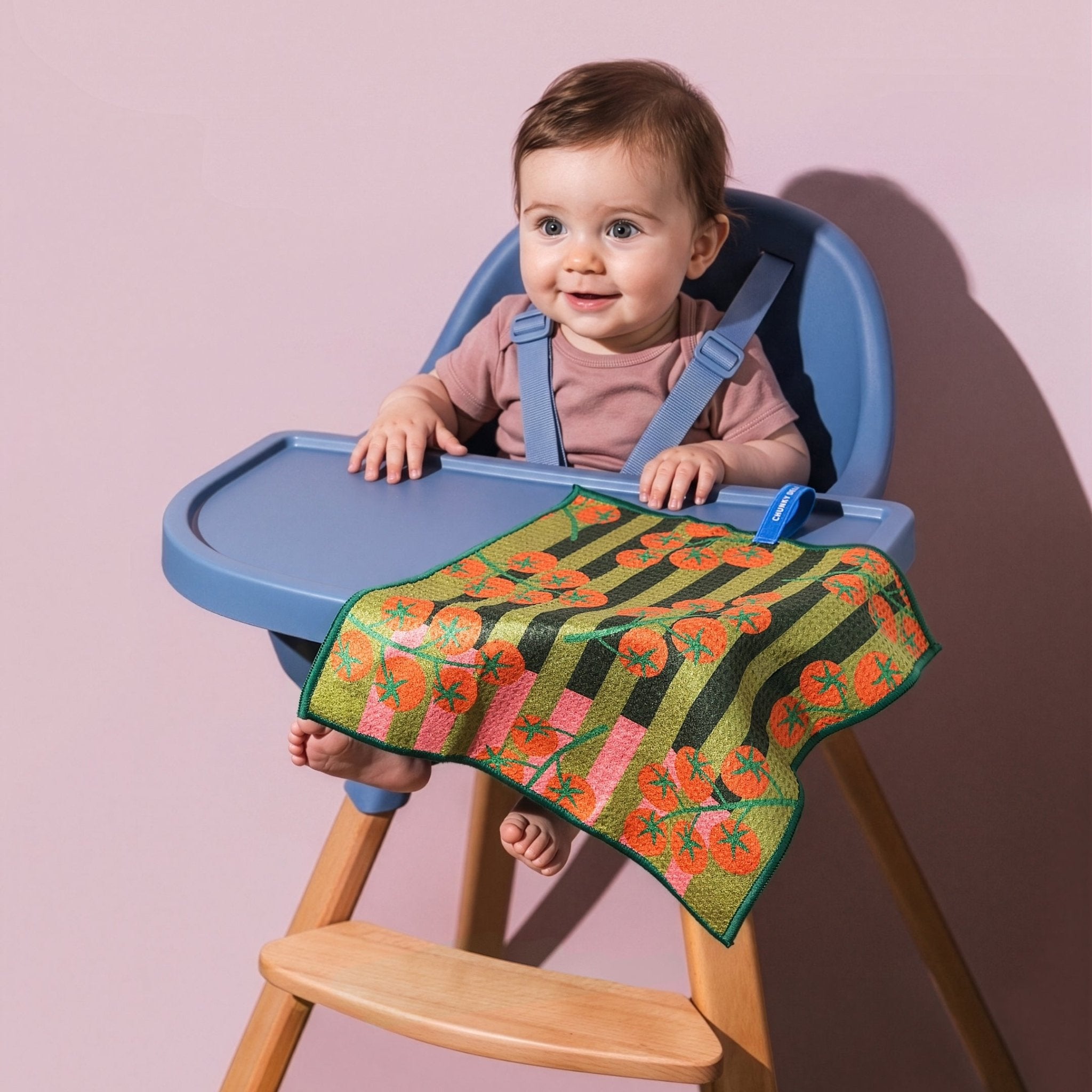 Chunky Deli product image of a Blue high chair holding a baby with a colorful striped tomato towel draped over it against a pink background