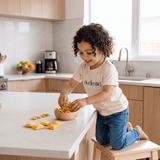 Child playing with felt pasta on a kitchen counter, wearing Al Dente Kids' Tee from Chunky Deli.