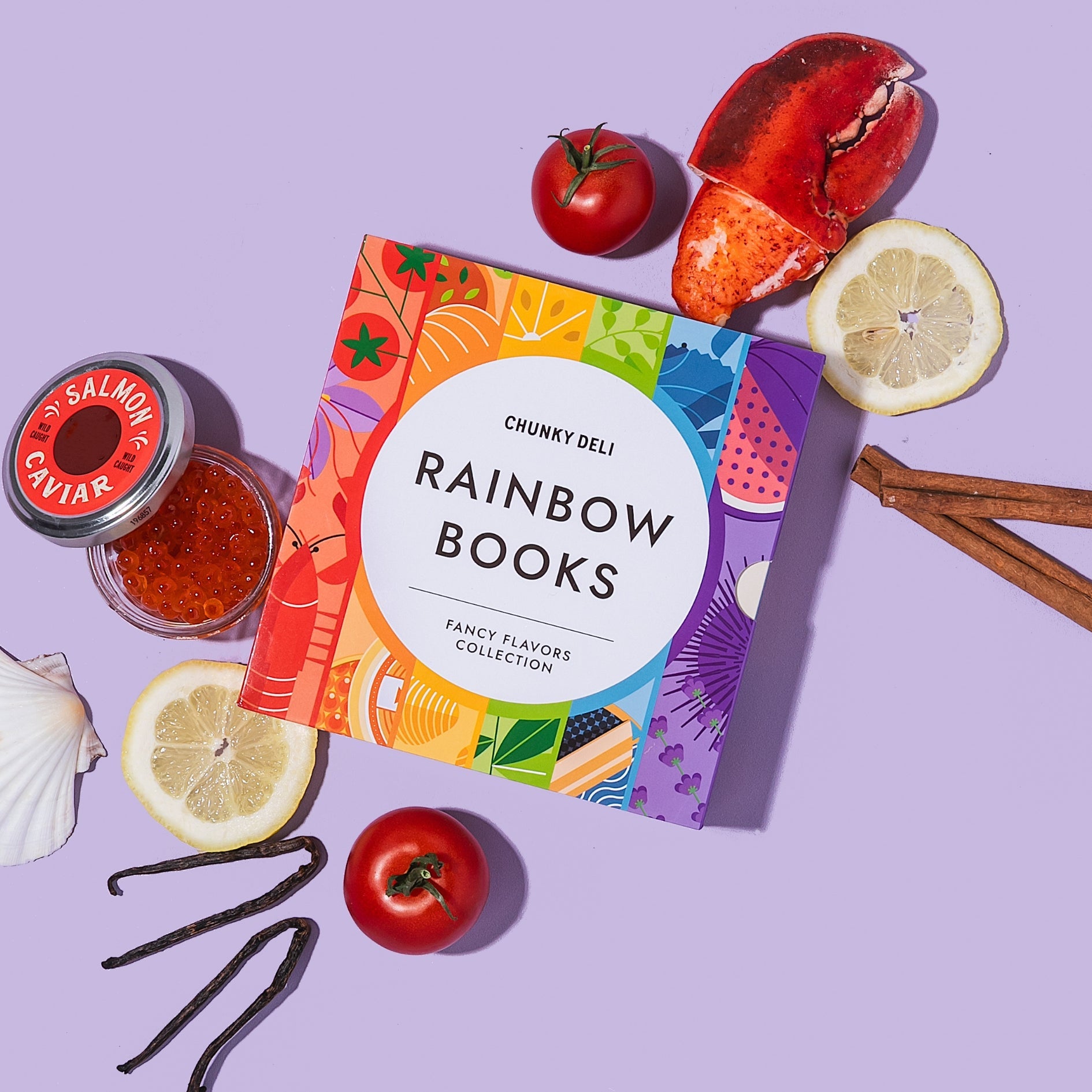 Chunky Deli "Rainbow Books Fancy Flavors Collection" slipcase, designed with a vibrant rainbow of food illustrations, surrounded by various food-related props including a lobster claw, salmon caviar, sliced lemons, tomatoes, vanilla beans, cinnamon sticks
