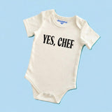 Chunky Deli product image: Natural-colored "Yes, Chef!" bodysuit (100% cotton) laid flat on a pastel blue background, showcasing the envelope shoulders and three-snap closure.